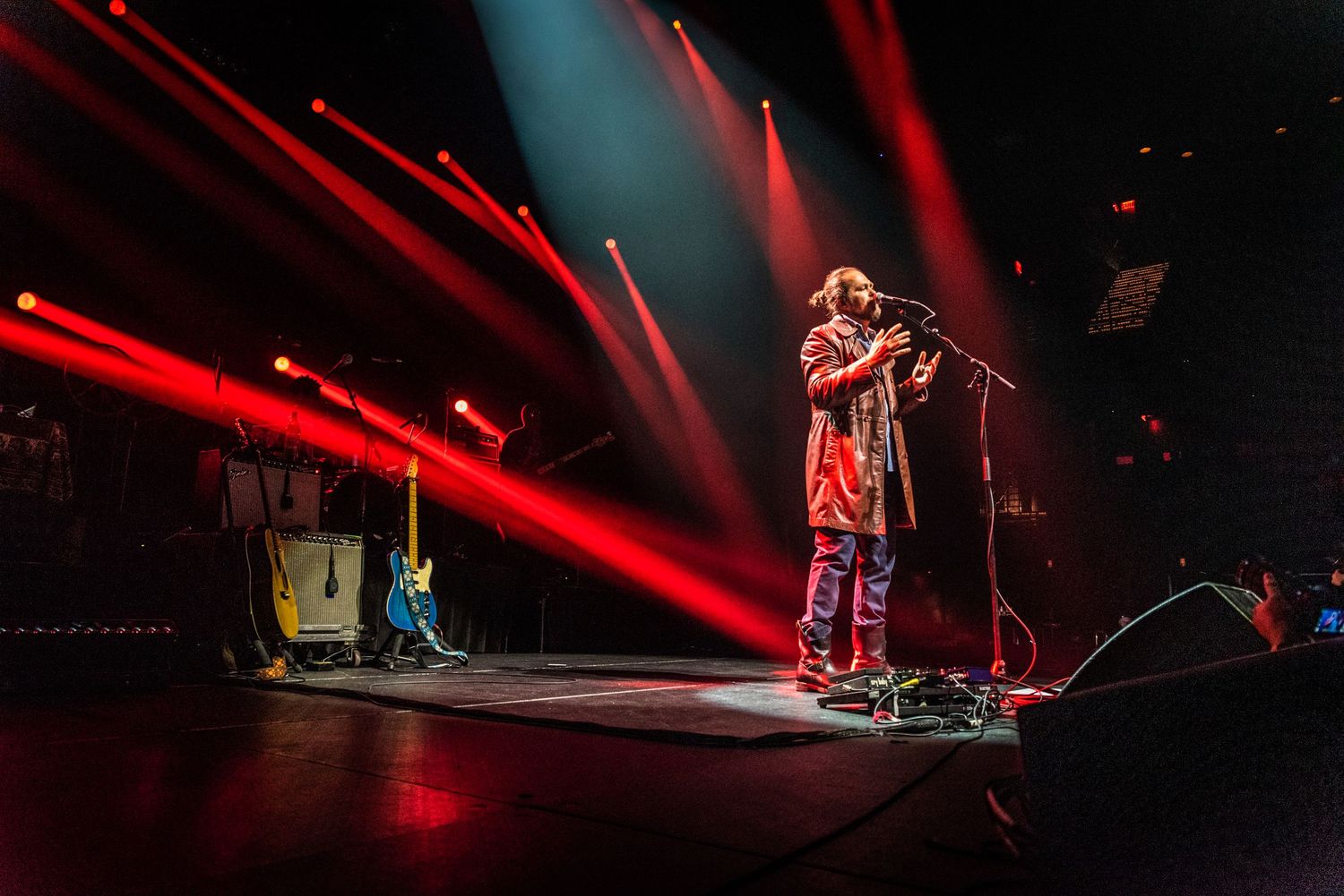 Solo performer stands in spotlight with red beams crossing overhead on dark stage.