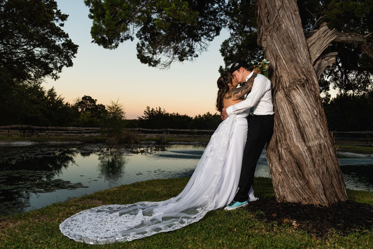Wedding couple embracing by tree at sunset with long white dress train.