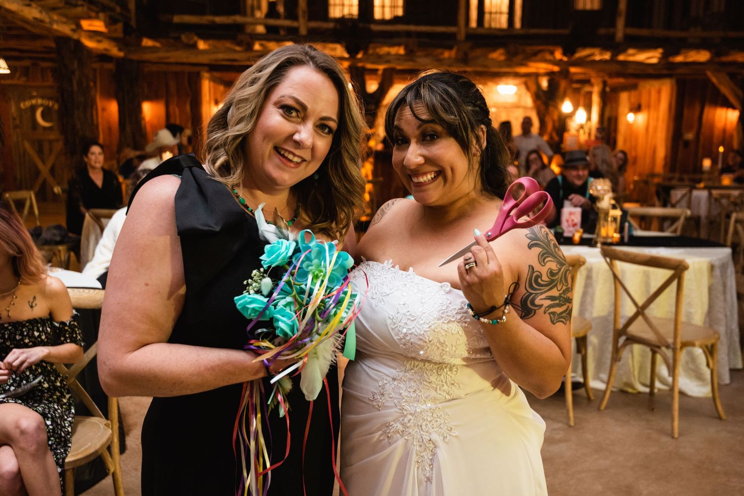 Two wedding attendees sharing a joyful moment while holding colorful bouquets in a rustic venue.