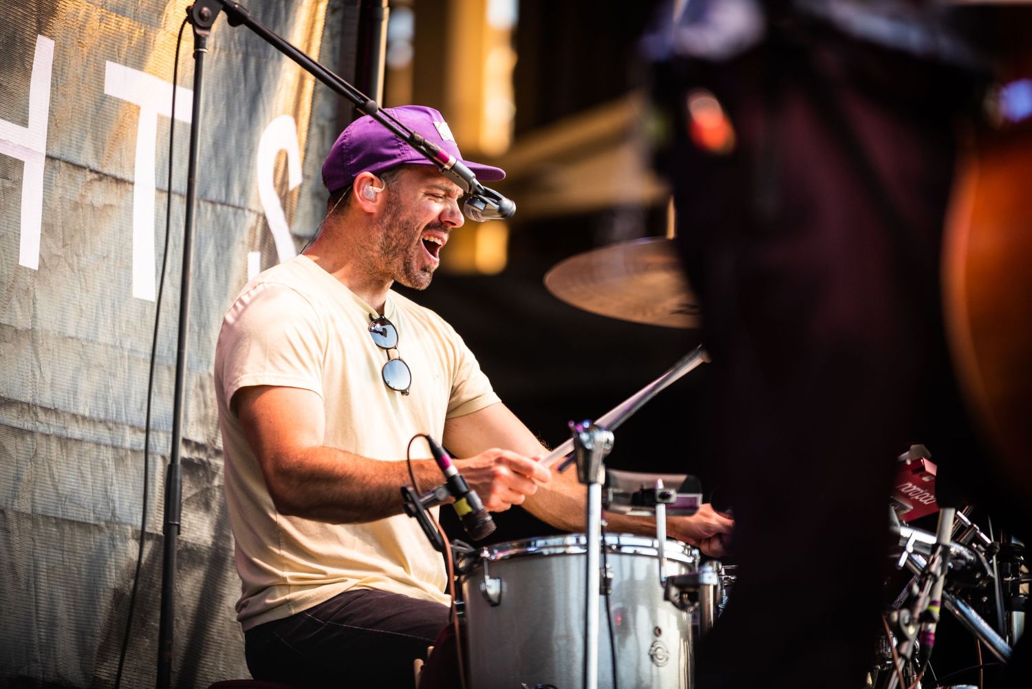 Drummer in purple cap performs enthusiastically behind drum kit on stage.