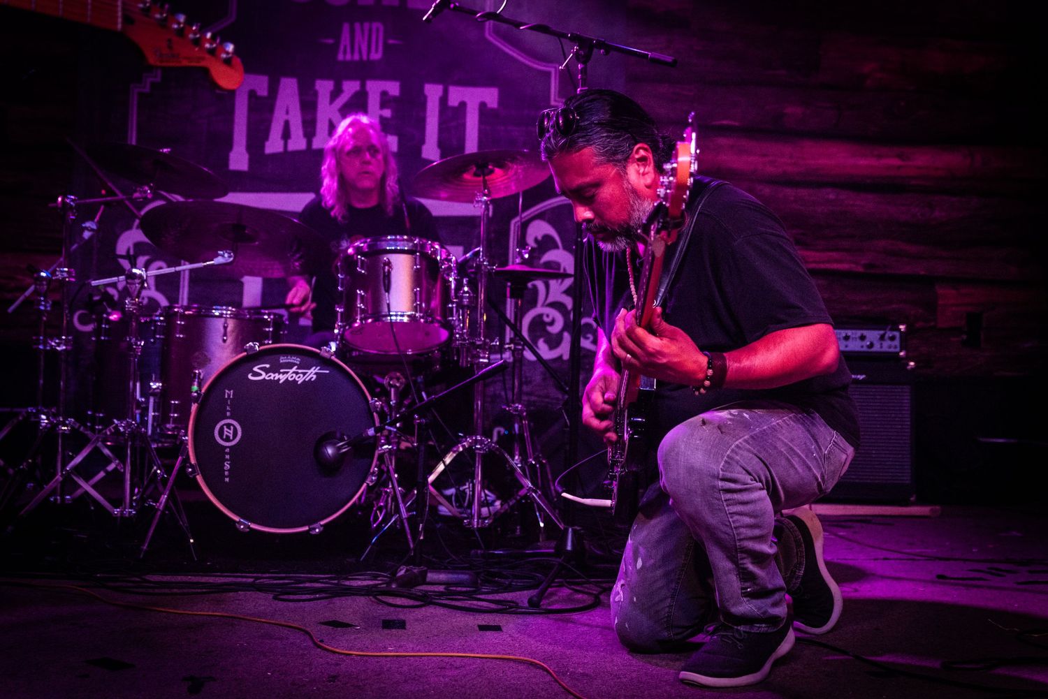 Musician performs on stage with black drum kit and purple lighting during a live concert.