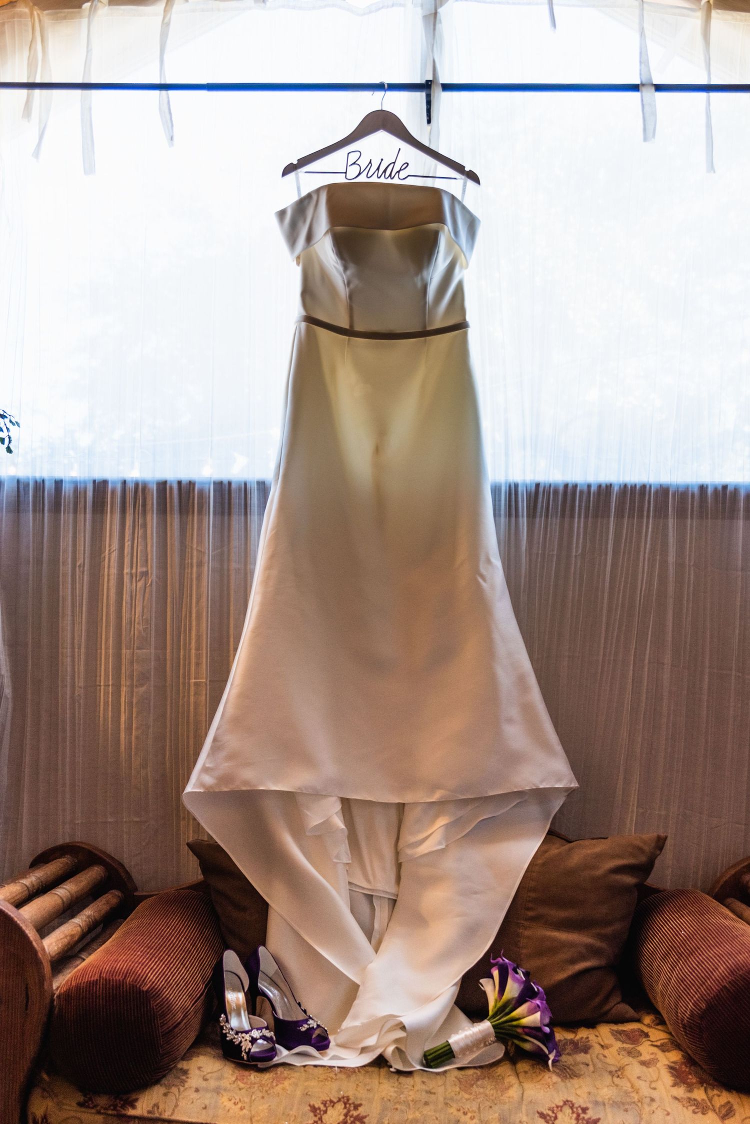 An elegant white wedding dress hangs on display against a brown curtained backdrop.