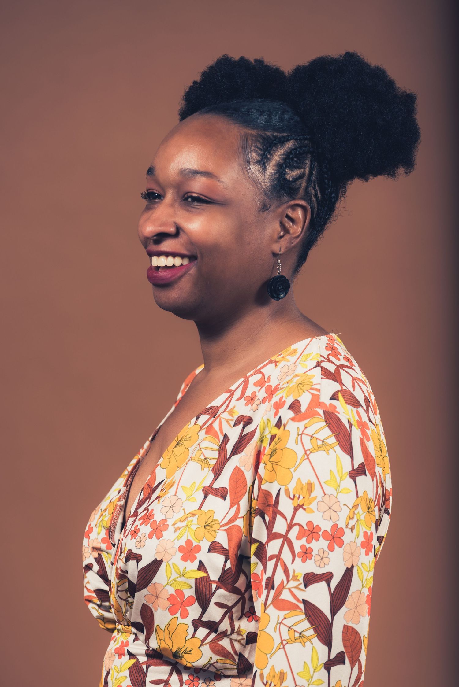 Side profile portrait showing intricate braided hairstyle and floral dress against brown background.