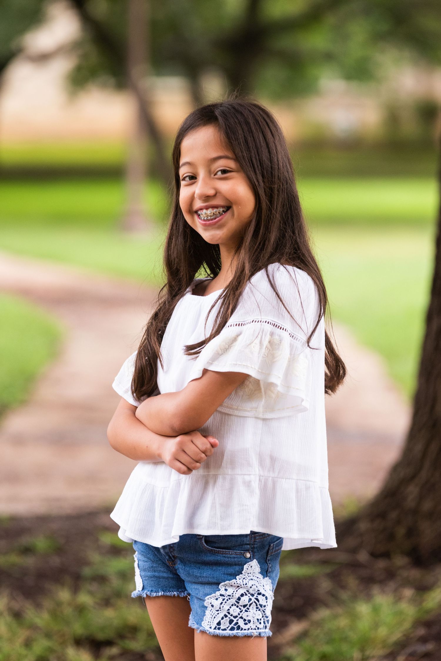 A young person in a white ruffled top and denim shorts smiles at the camera outdoors.