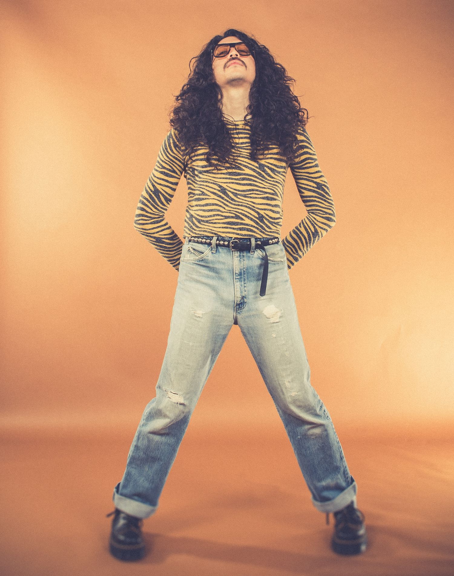 Person in tiger print top and ripped jeans poses energetically against orange background.