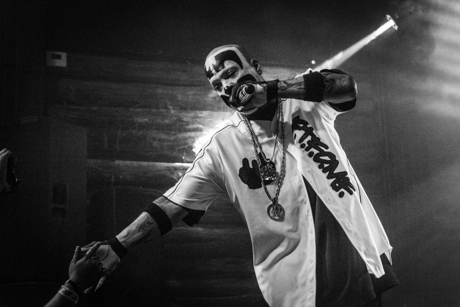 Stage performer in white clothing with face paint performing energetically under dramatic lighting.