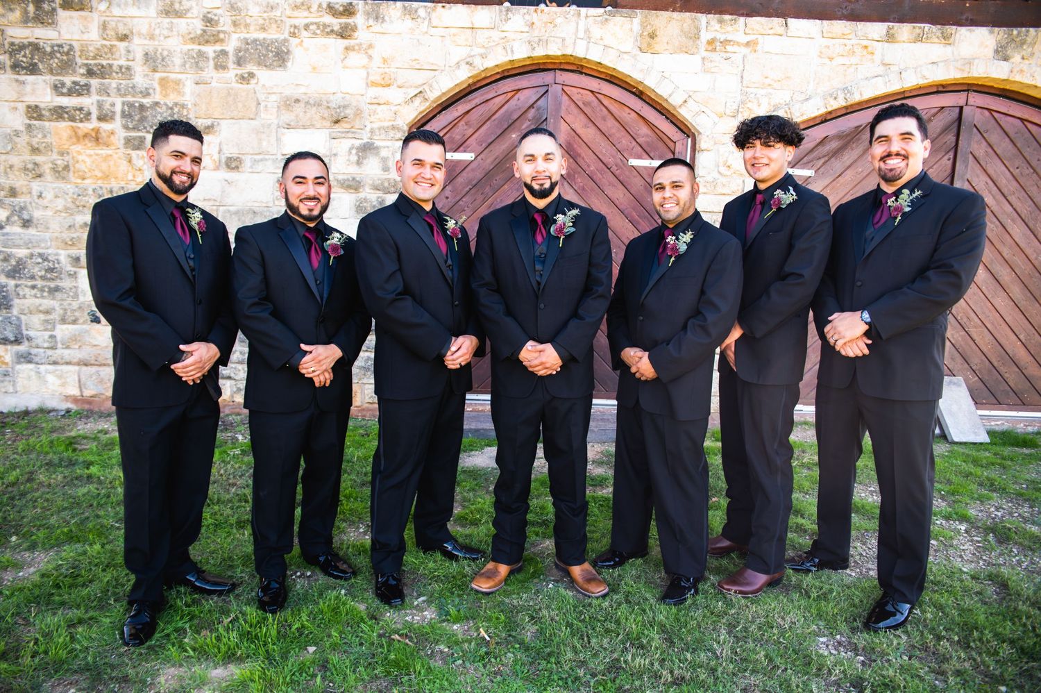Groomsmen in black suits and red ties pose together against rustic wooden doors and stone wall backdrop.