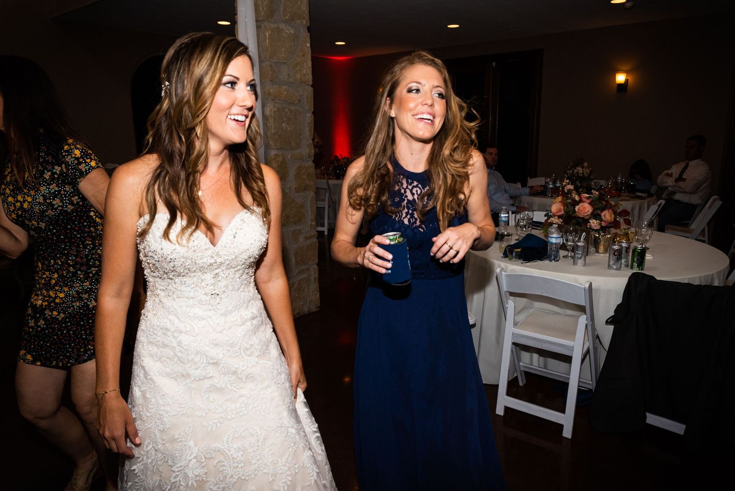 Two women in formal attire share laughter while walking through a dimly lit reception venue.