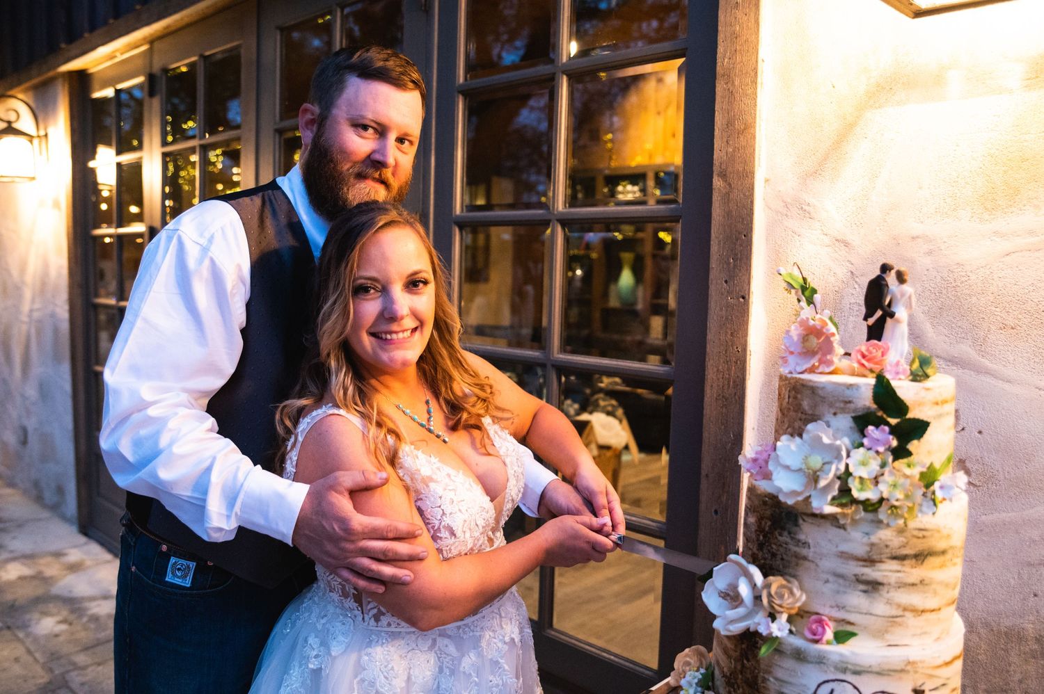 Newlyweds pose beside their elegant white wedding cake with floral decorations.