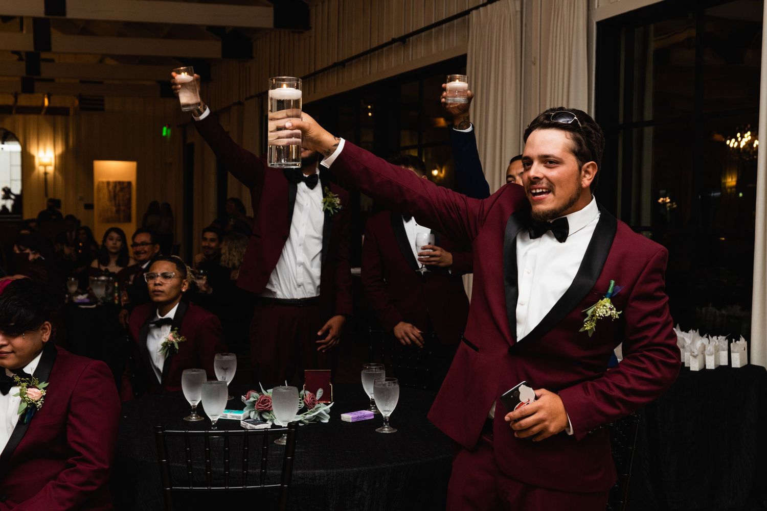 A celebration toast in a dimly lit reception venue with burgundy tuxedos.