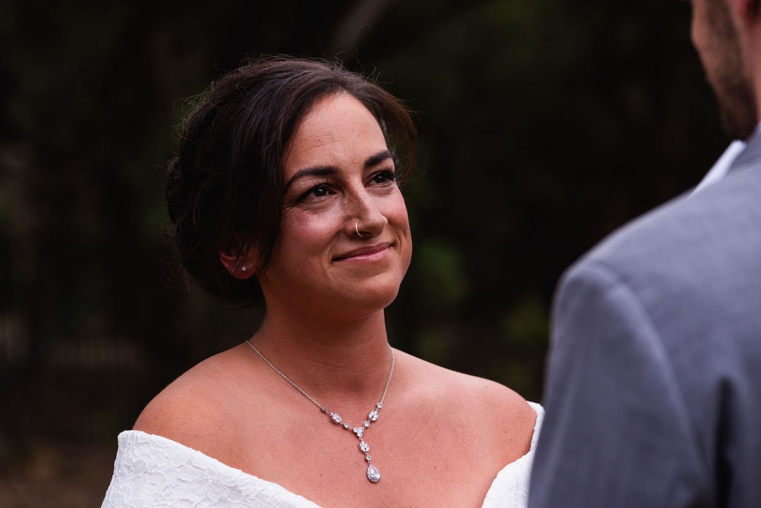 Bride in off-shoulder white dress smiles radiantly while wearing elegant crystal necklace.