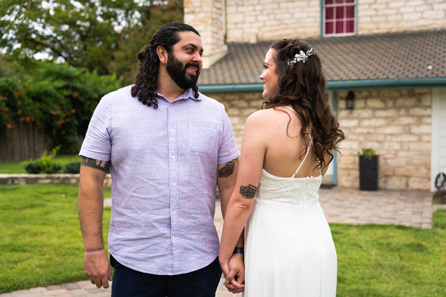A couple shares an intimate moment in front of a brick building with a garden backdrop.