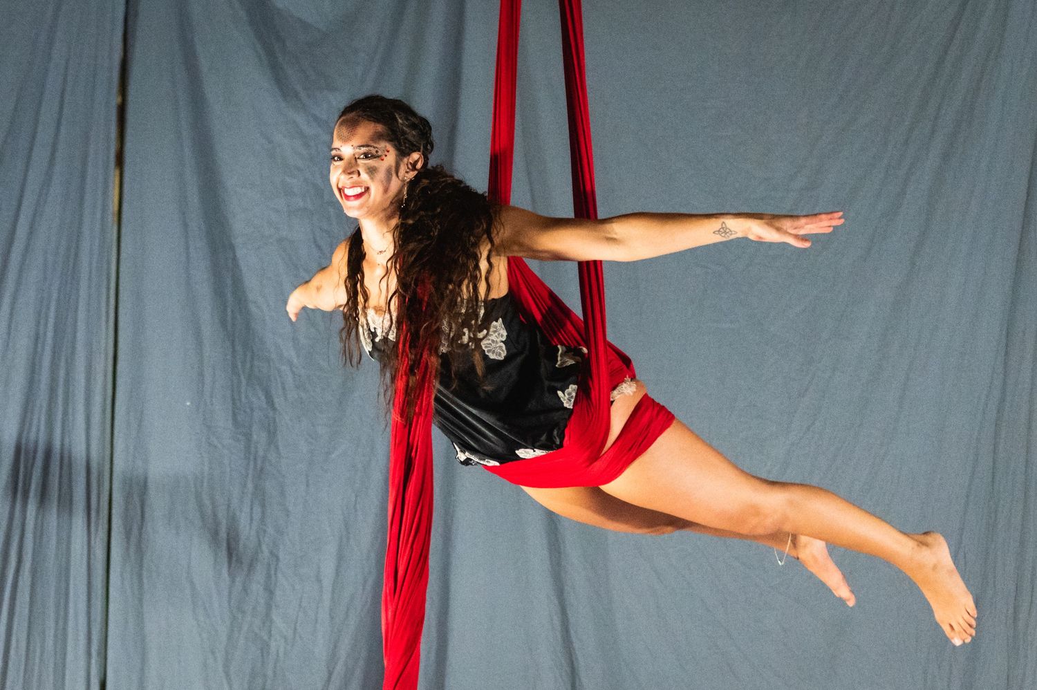 Aerial artist performs a flying pose while suspended from red silks in a studio setting.