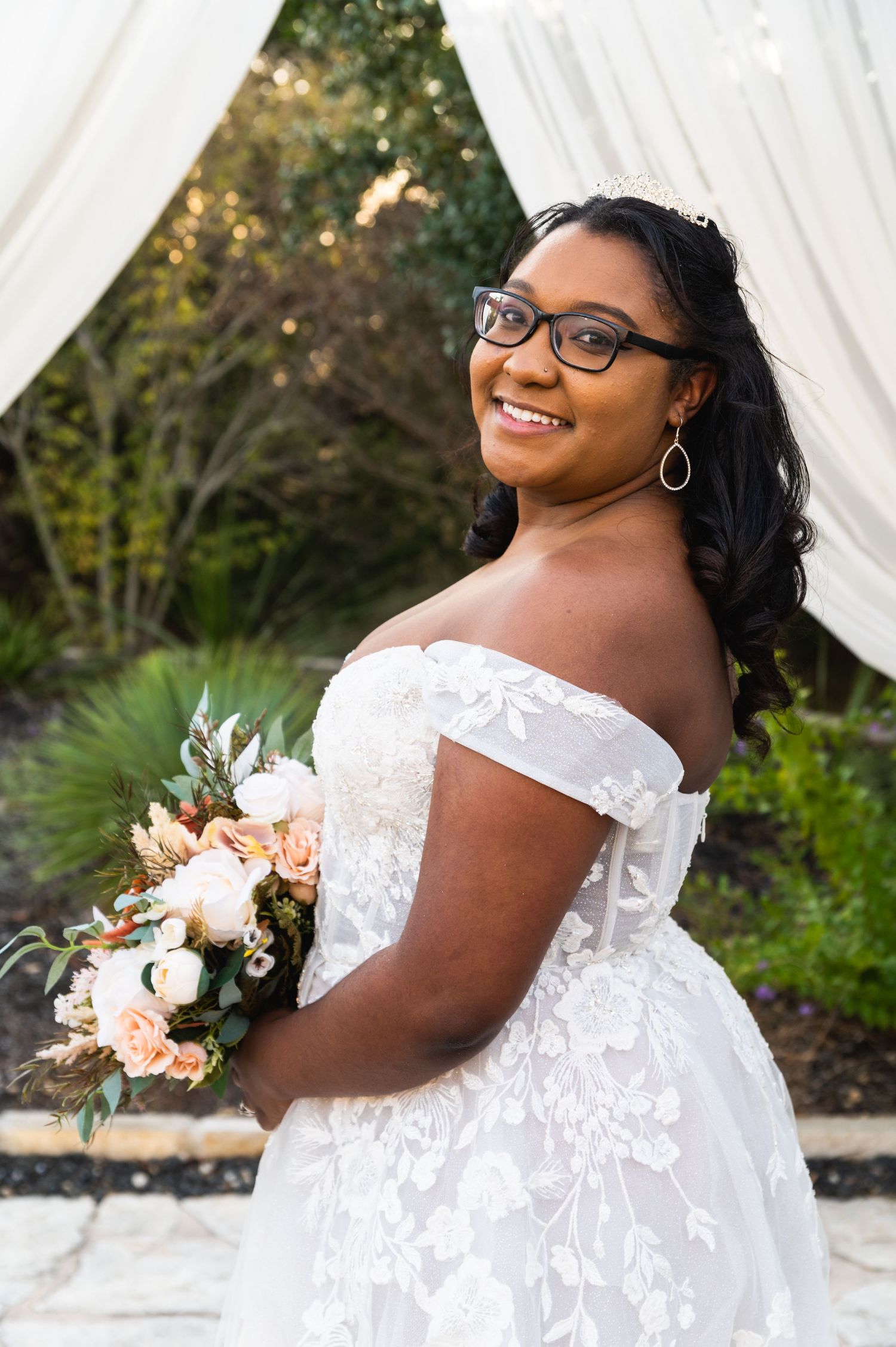 A bride in a white off-shoulder lace gown holds a peach and white bouquet while smiling.