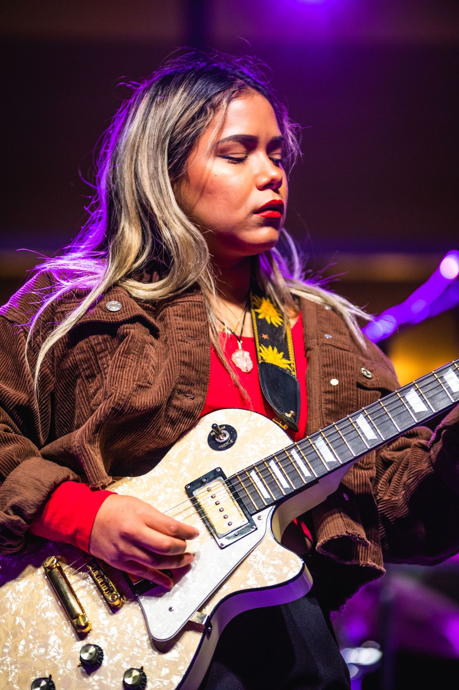 Musician plays white electric guitar in brown jacket under purple stage lighting.