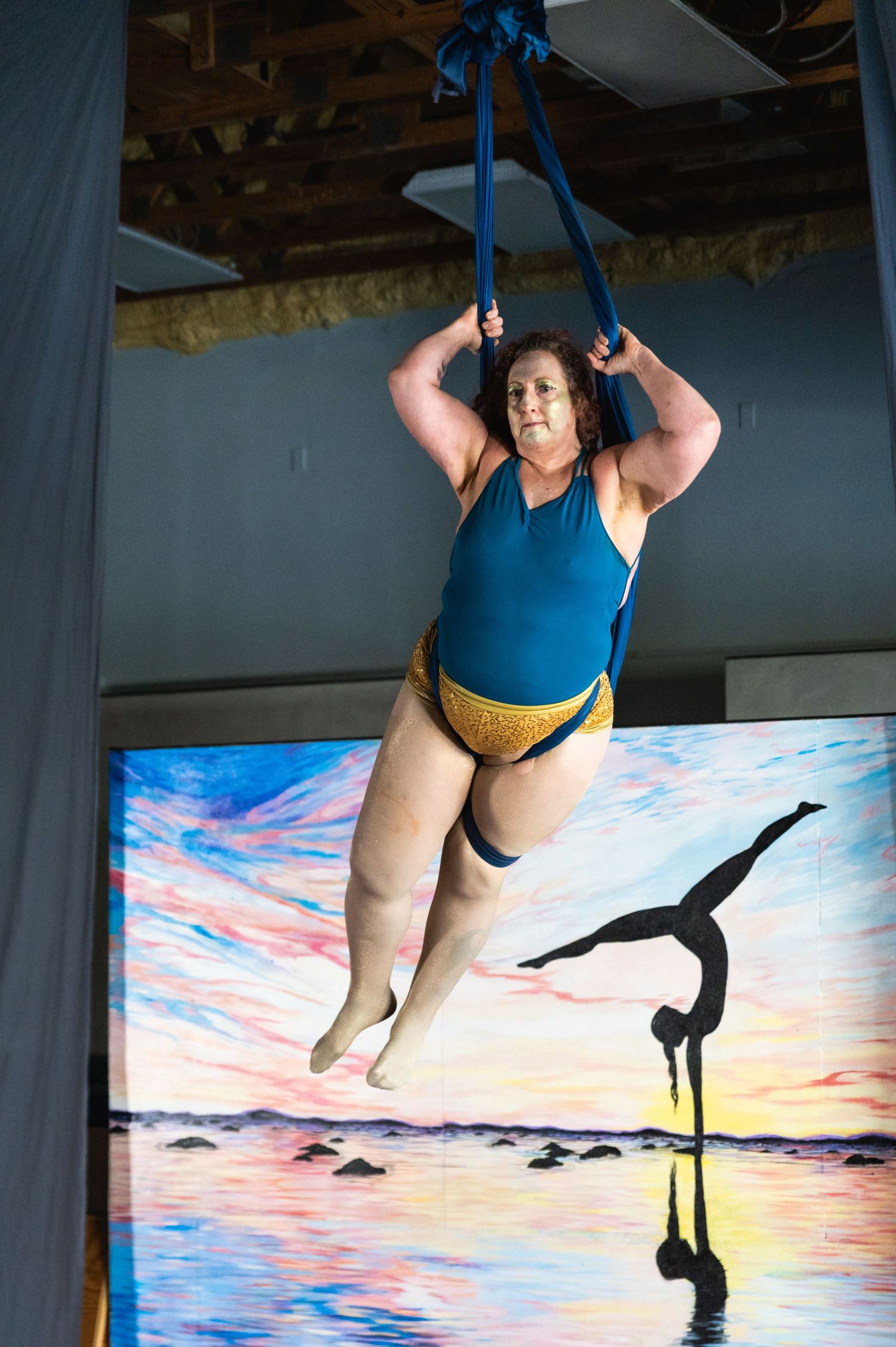 Aerial artist in blue and gold leotard performs suspended against sunset backdrop.