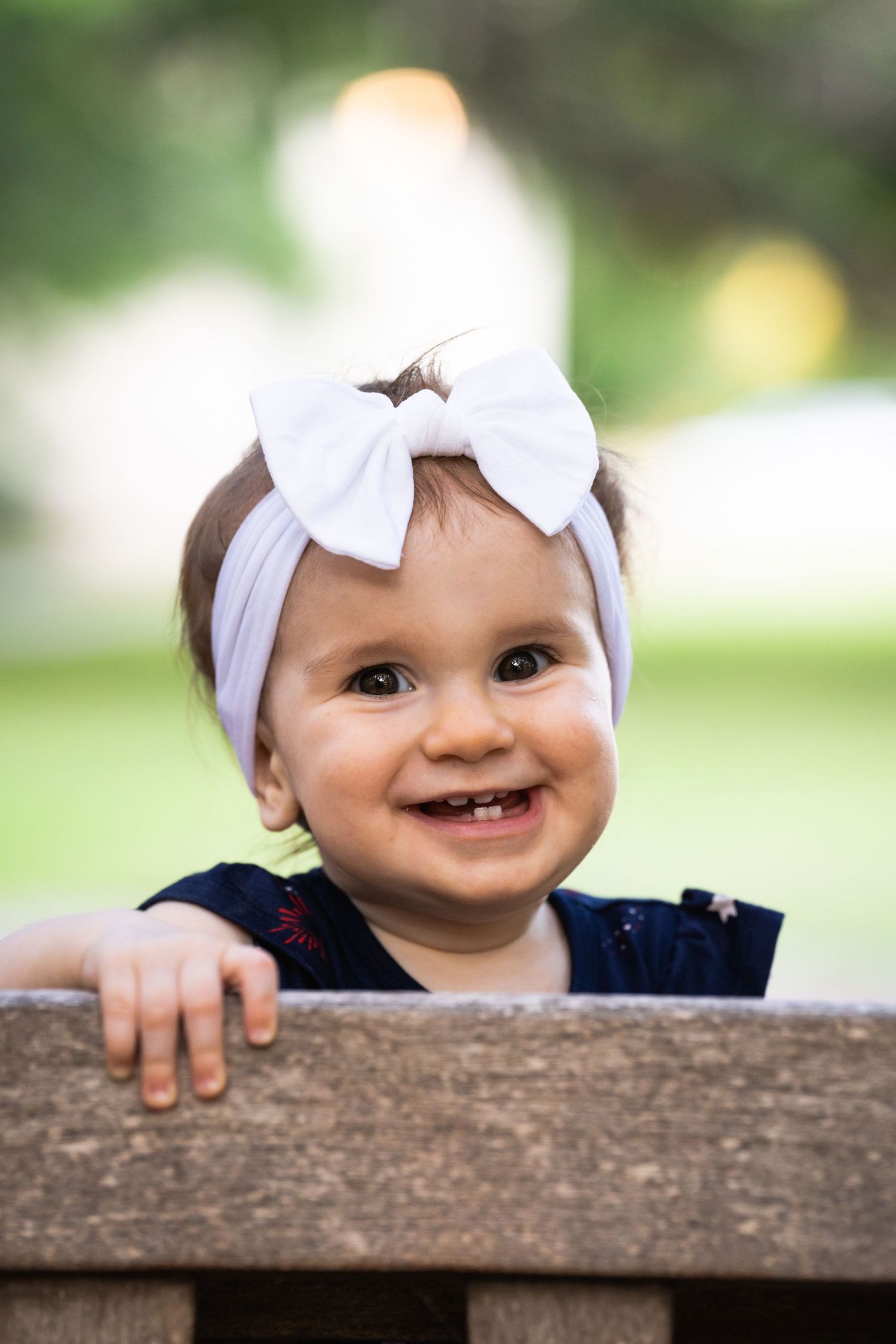 Baby in navy dress and white bow headband peeks over wooden railing outdoors.