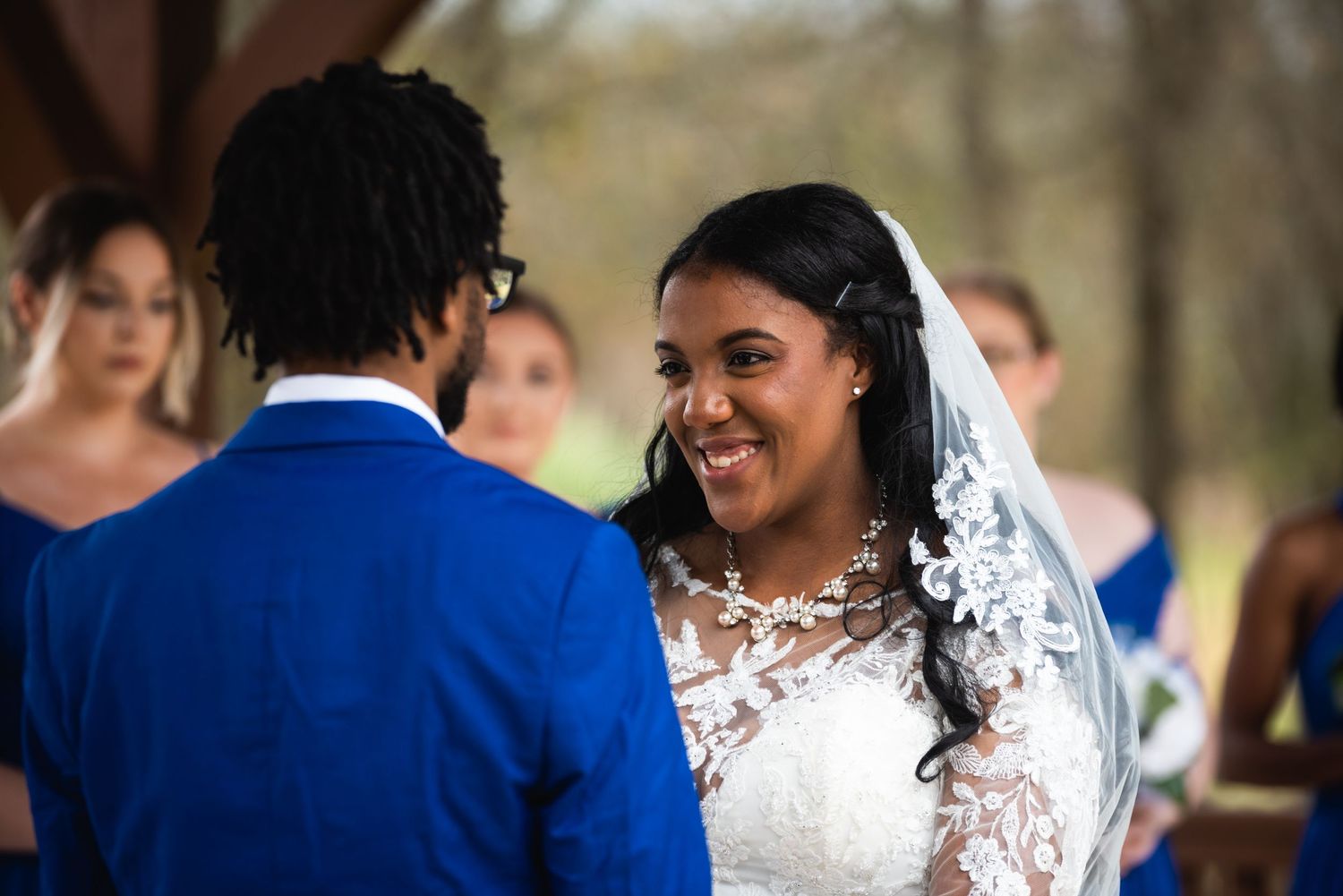 Bride in a white lace dress smiles joyfully during wedding ceremony alongside groom in royal blue suit.