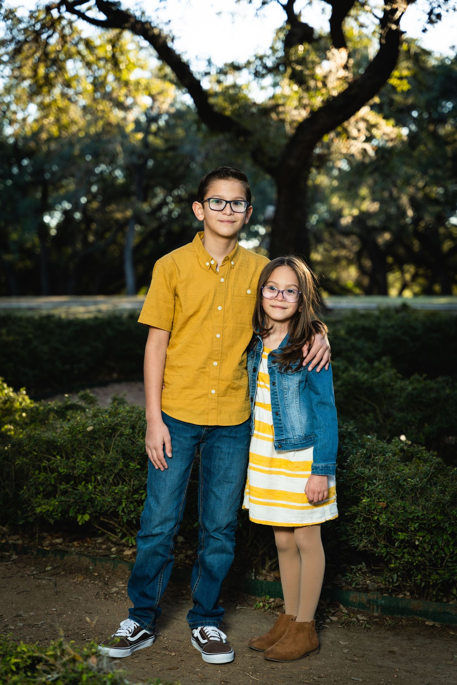 Two siblings stand together on a path with trees in the background during golden hour.