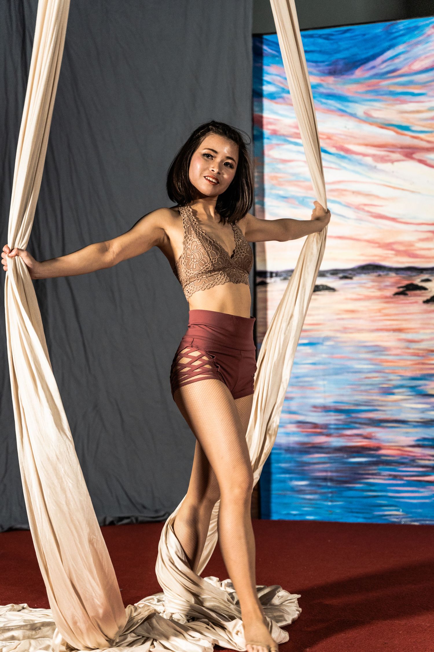 Aerial silk performer poses gracefully against a painted sunset backdrop in a dance studio setting.