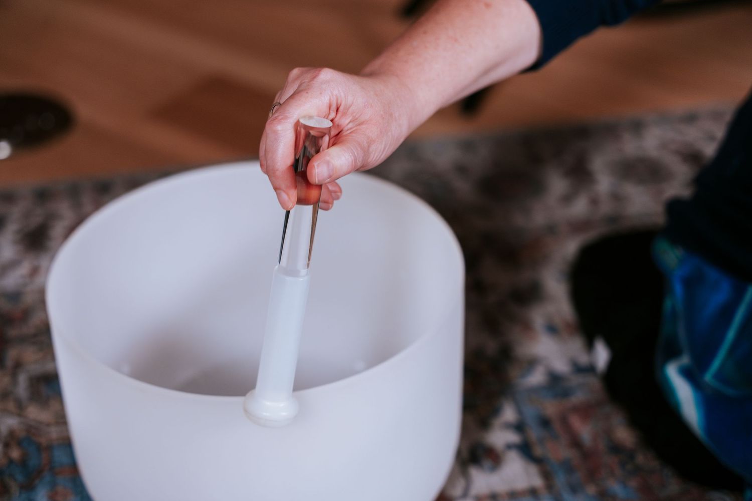 A glass mixing bowl pours liquid into a white porcelain bowl on a wooden counter while someone adds ingredients.