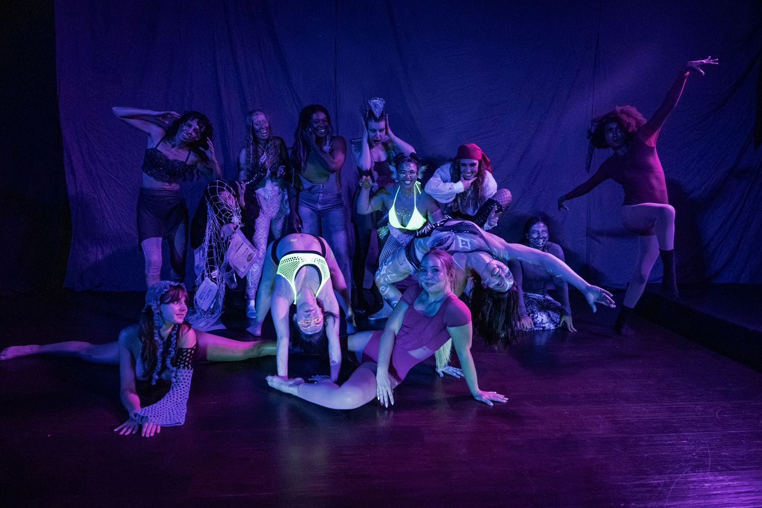 Dancers perform a dramatic group pose under purple stage lighting against a dark backdrop.