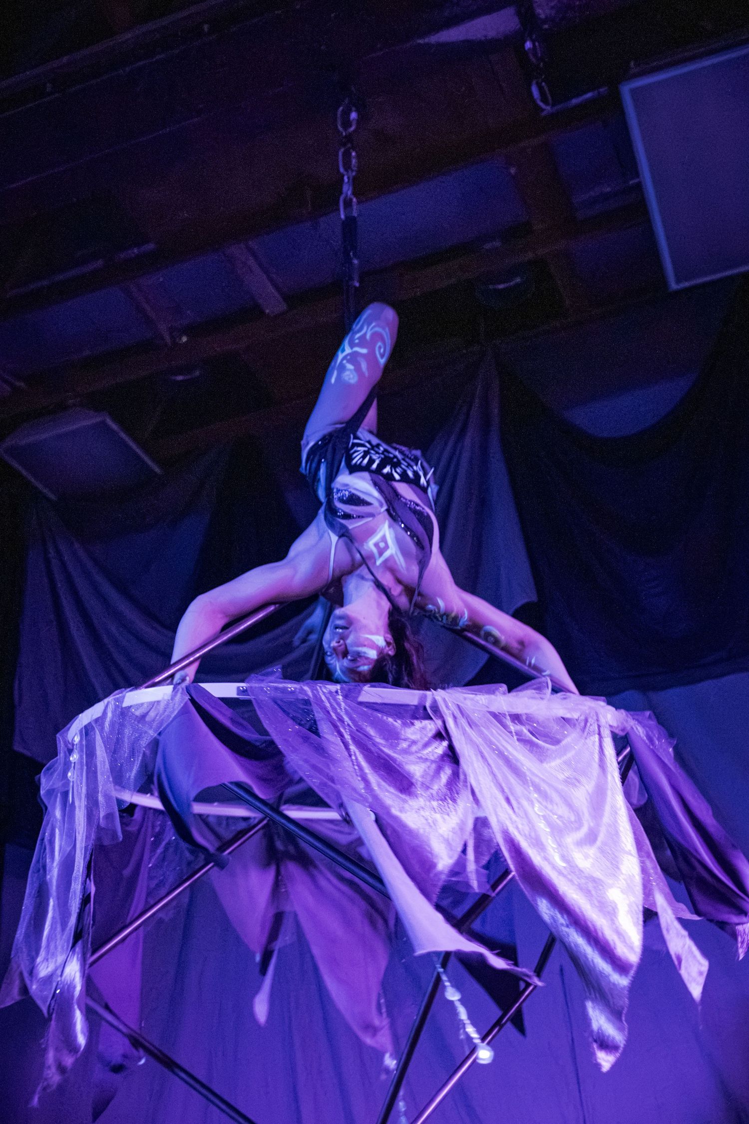 Aerial acrobat performs inverted pose on suspended apparatus under purple stage lighting.