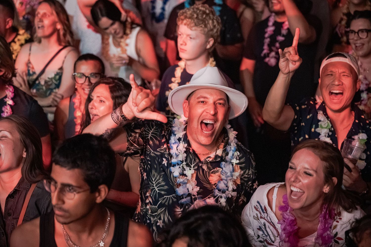 Excited crowd at a tropical-themed concert, with people wearing leis and Hawaiian shirts, cheering enthusiastically.