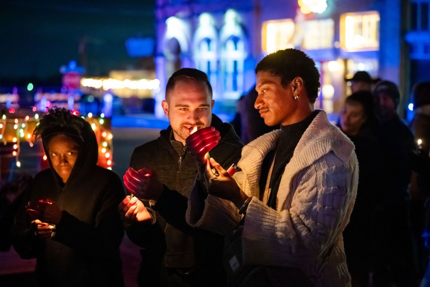 People lighting cigarettes at night in a dark urban setting with glowing lights in the background.