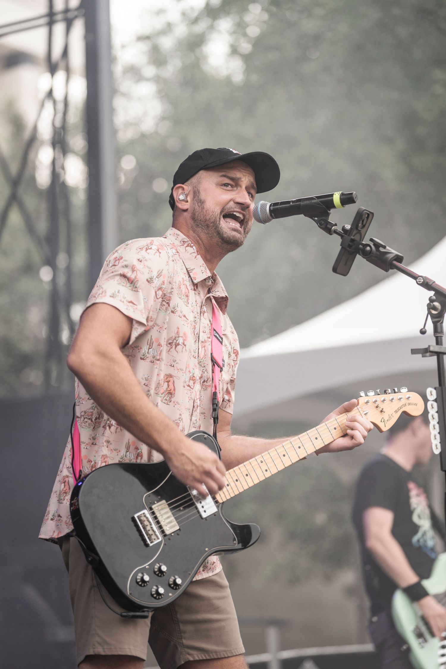 Musician performs energetically on stage with a black electric guitar while wearing a floral print shirt and baseball cap.
