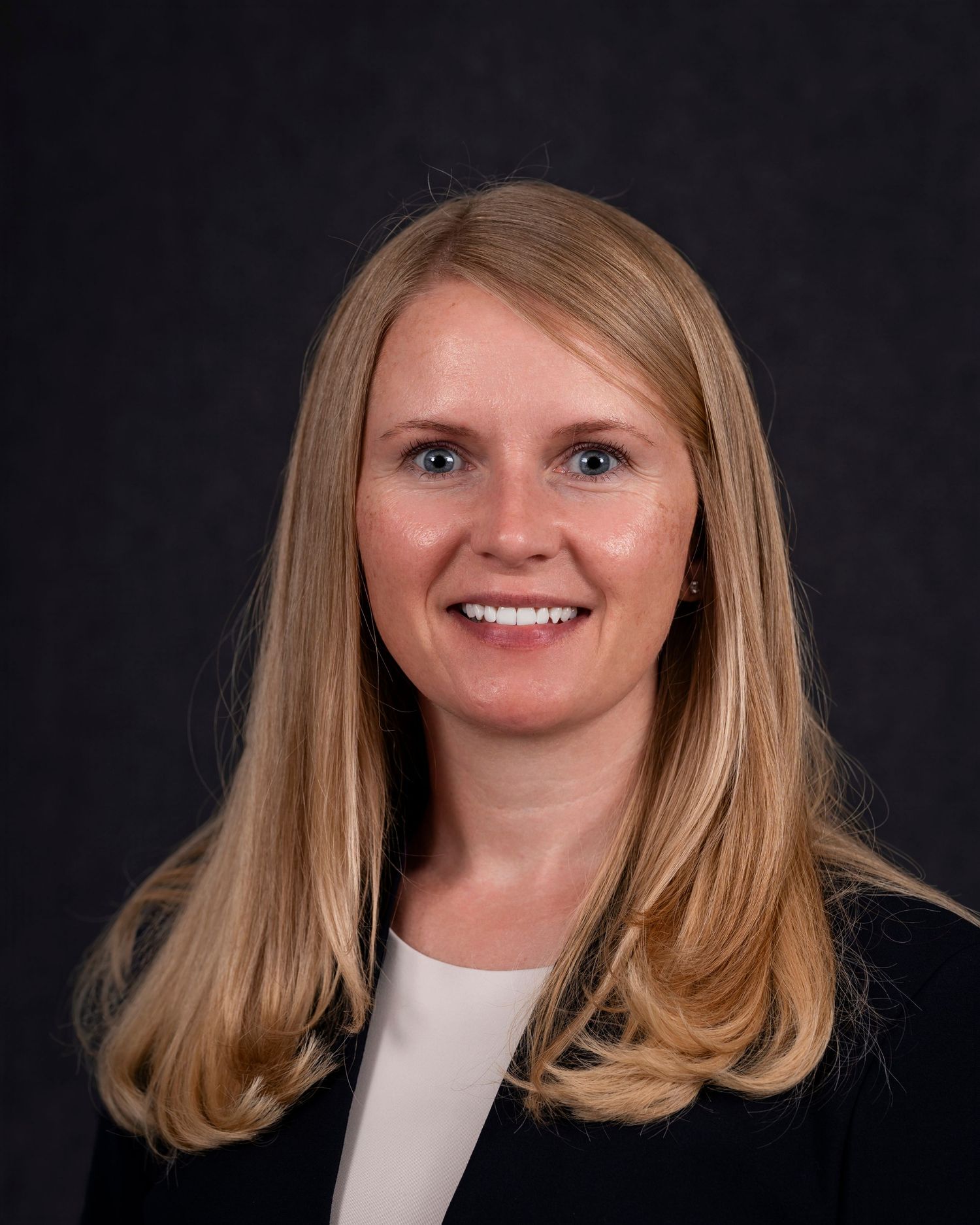 Professional headshot of someone with blonde hair wearing a black blazer and white top against a dark background.