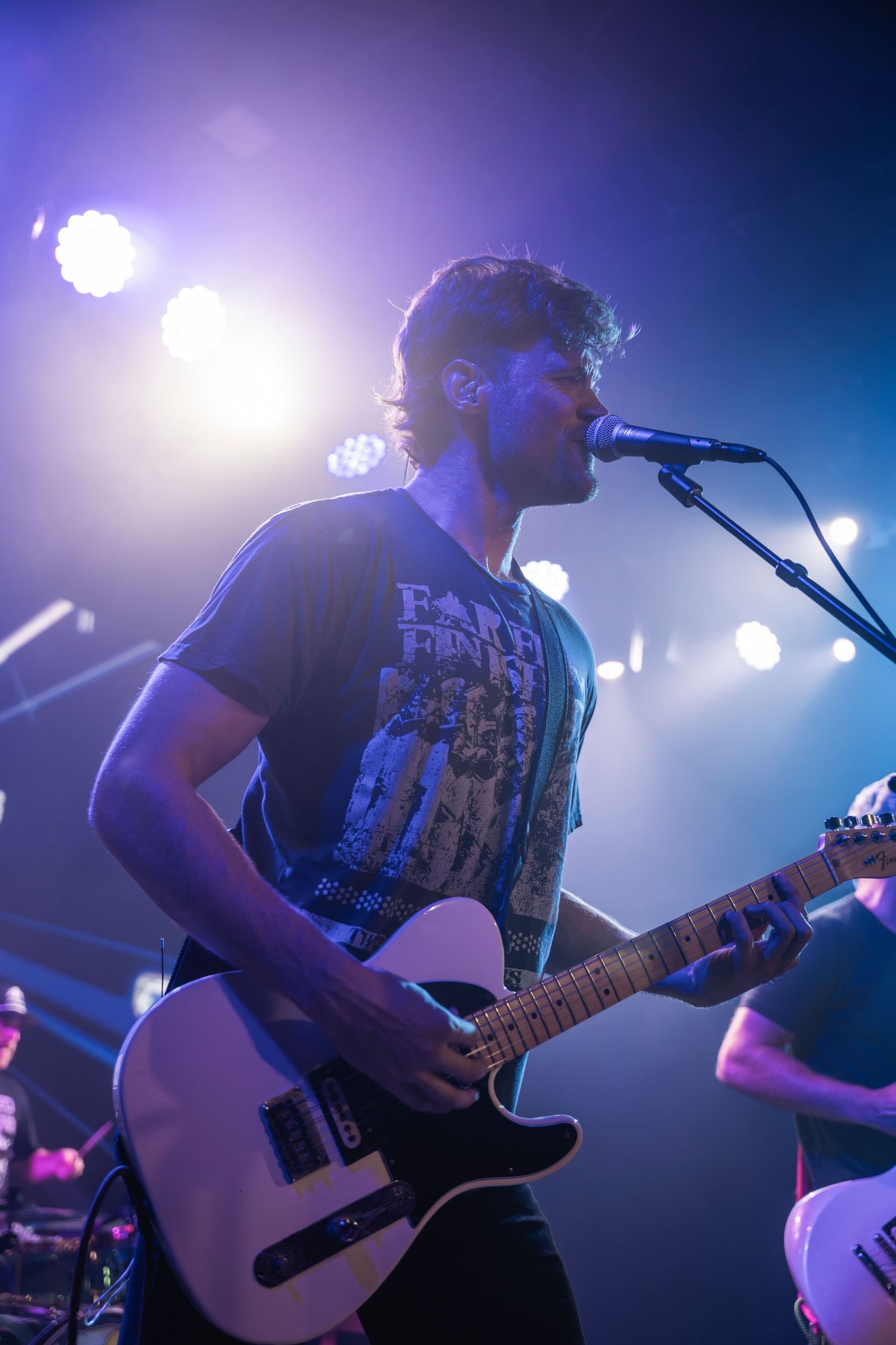 Musician performs on stage with white guitar under dramatic blue and purple concert lighting.