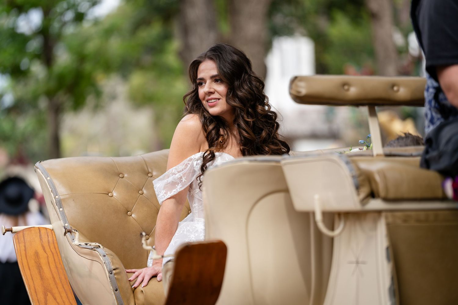 A person in a white off-shoulder dress sits on an elegant beige vintage sofa outdoors during a special event.