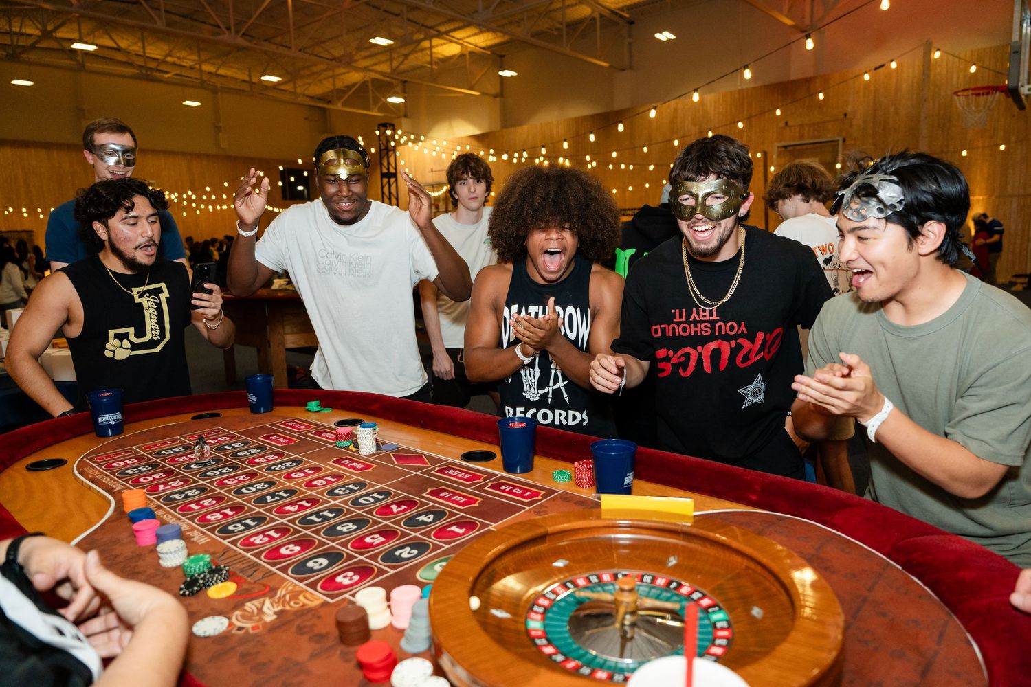 A group of excited players gather around a roulette table in a casino with warm lighting and festive atmosphere.