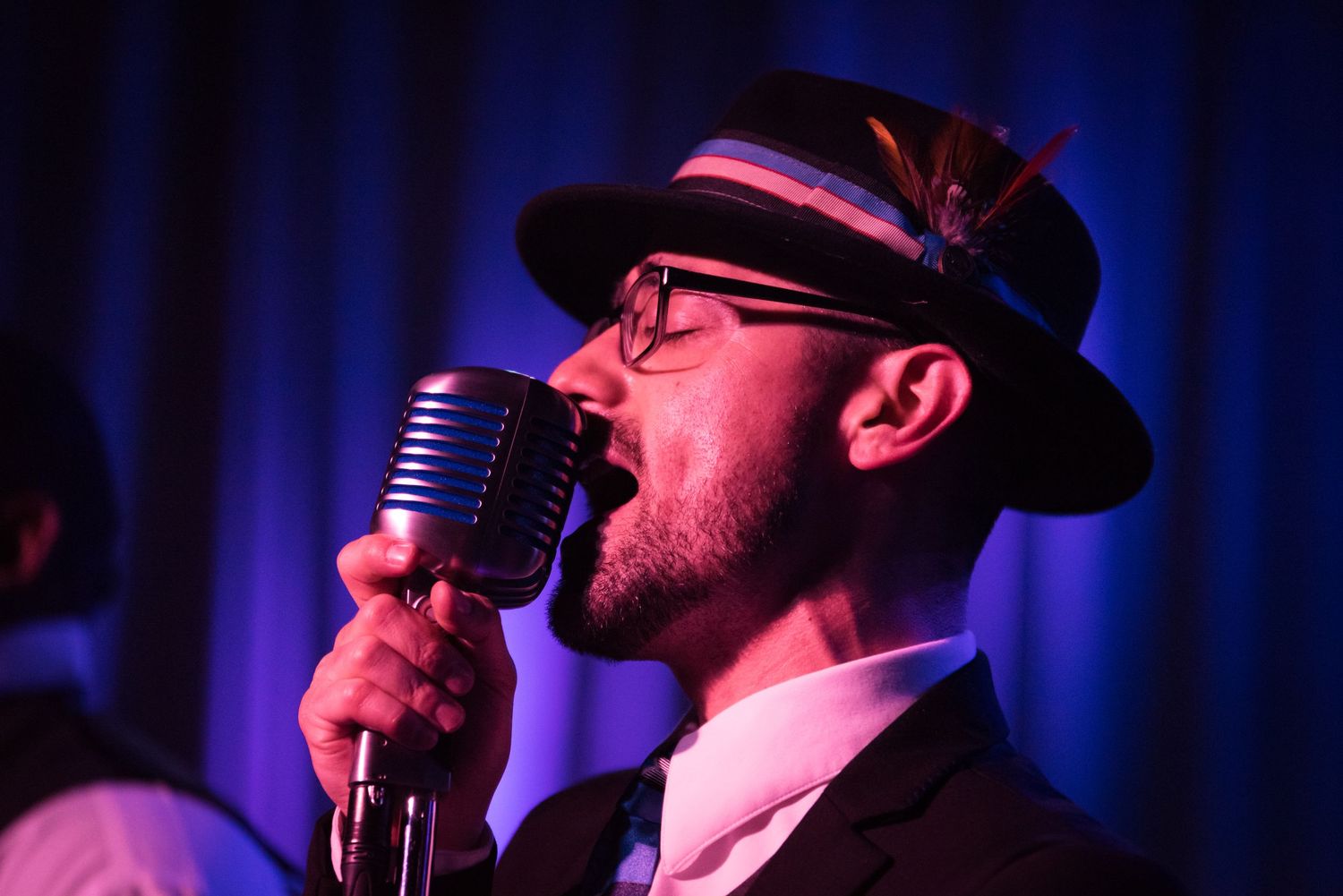 Performer in a black hat sings passionately into a microphone against a blue-lit stage backdrop.