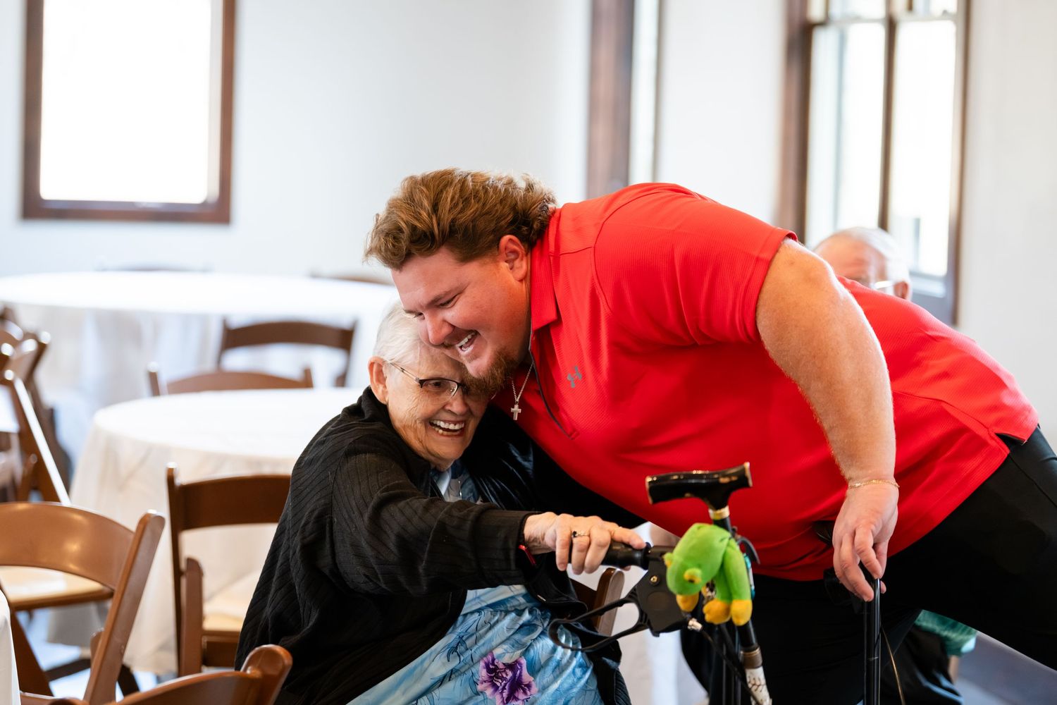 Caregiver in red shirt joyfully embraces elderly person in wheelchair at a bright indoor dining area.
