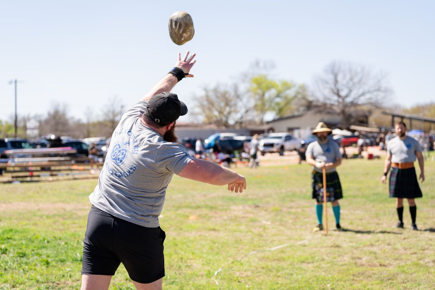 Man throwing a stone in a Highland Games competition on a sunny day with spectators in kilts watching.
