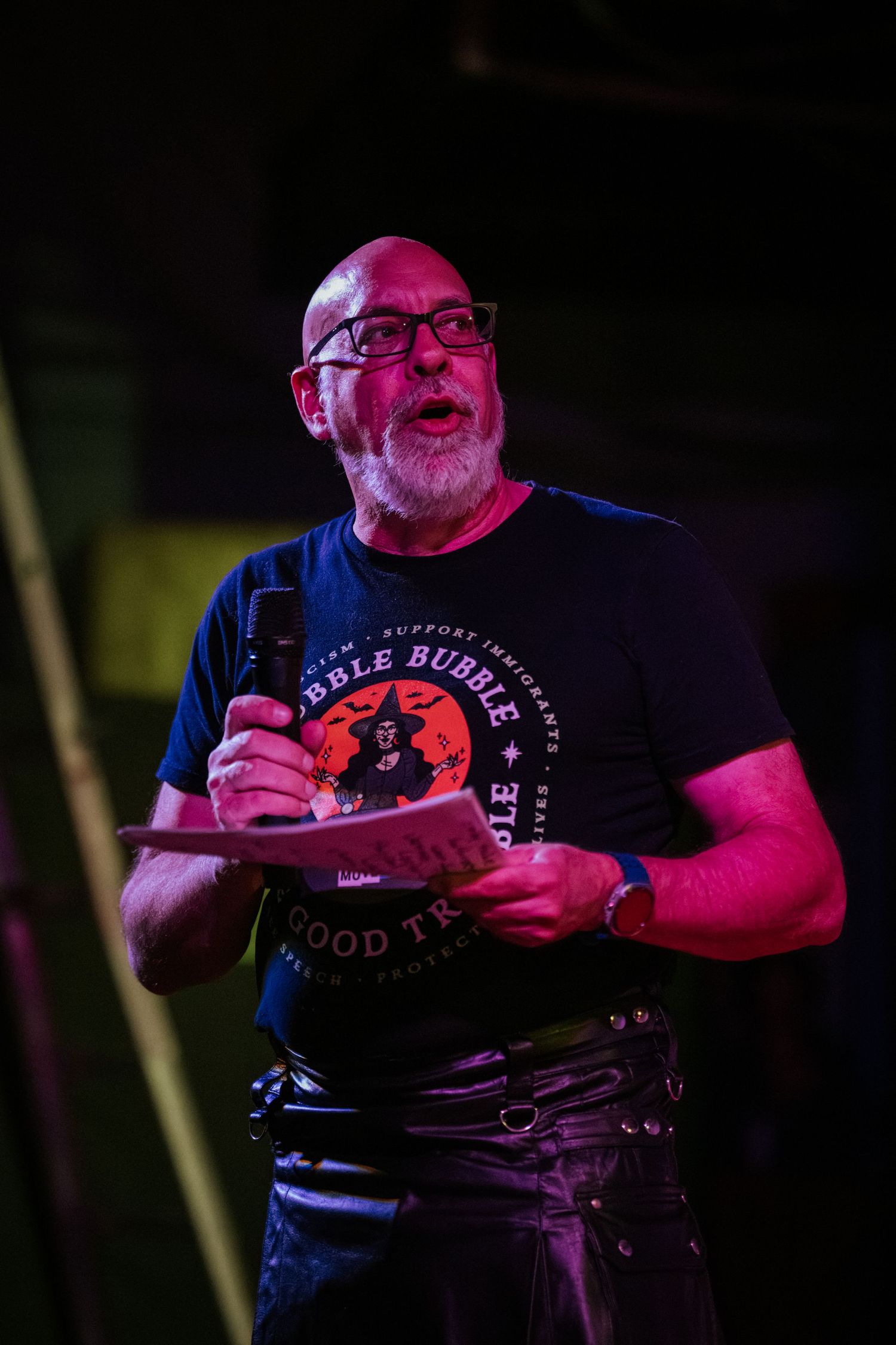 Bald man with glasses speaking into microphone on stage, wearing a dark graphic t-shirt under colorful pink stage lighting.