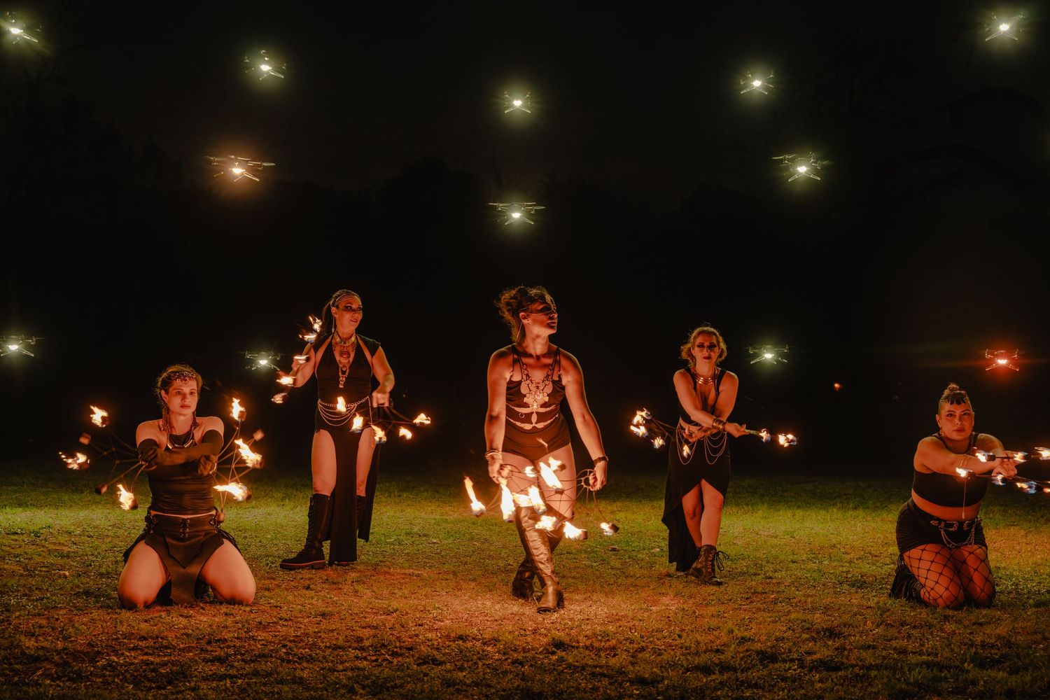 Five fire dancers perform at night, holding glowing flames in a dramatic outdoor display with bokeh lights in the background.