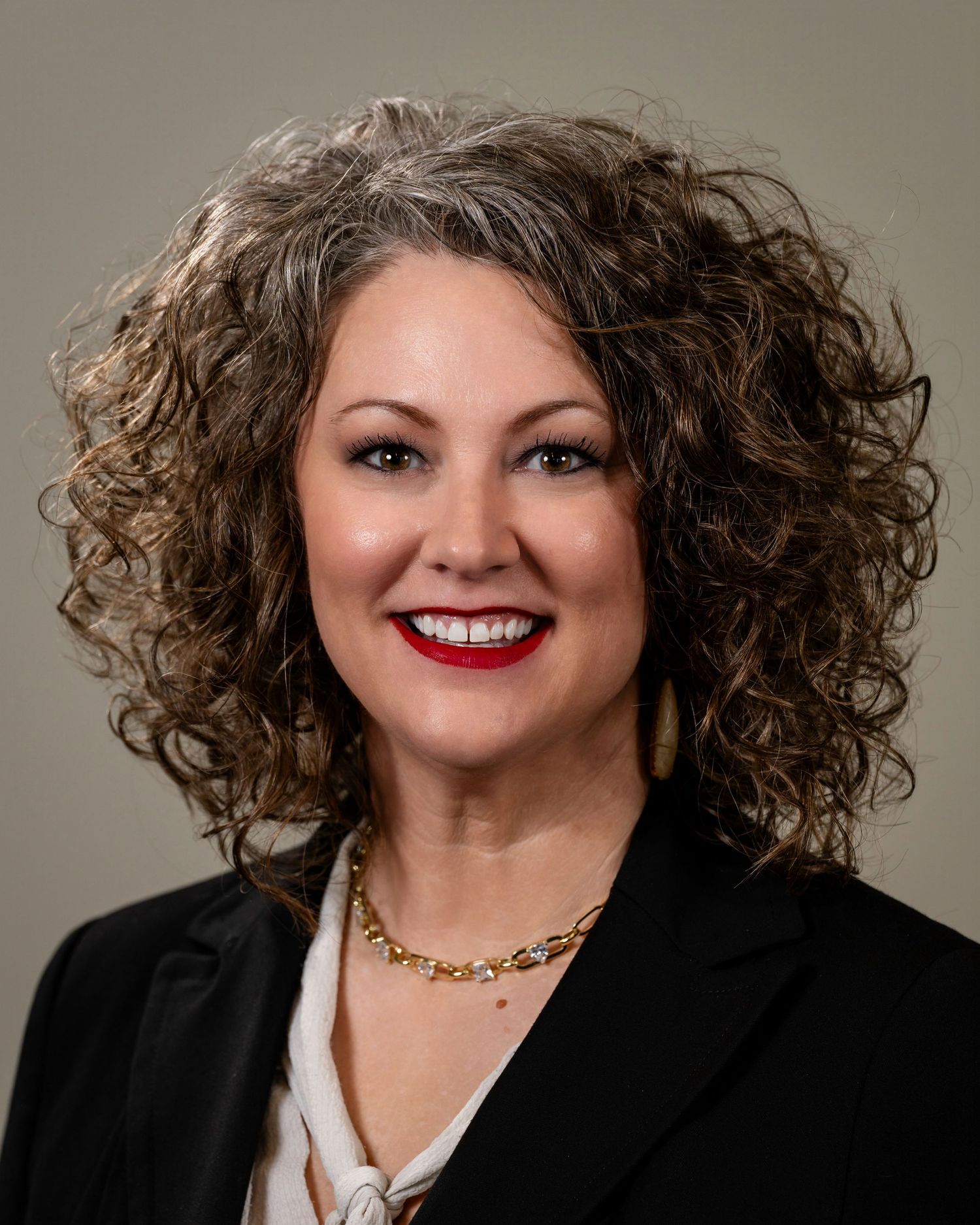 Professional headshot of a smiling woman with curly gray-brown hair, red lipstick, gold necklace, and black blazer.