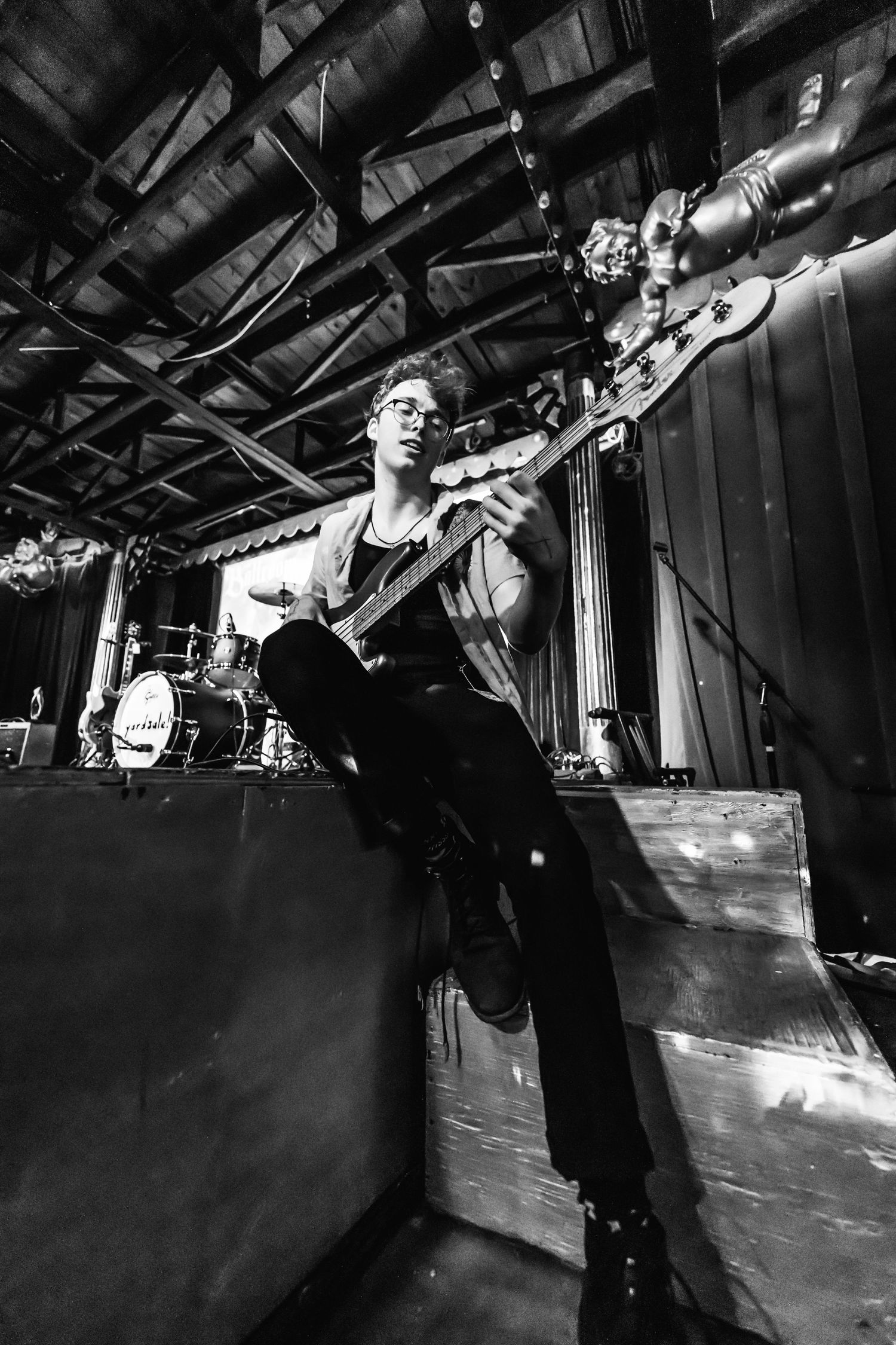 Black and white photo of musician playing guitar on stage with dramatic lighting and industrial ceiling.