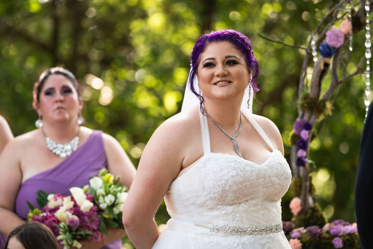 A bride wearing a white strapless dress and purple hair accessory smiles during an outdoor wedding ceremony.