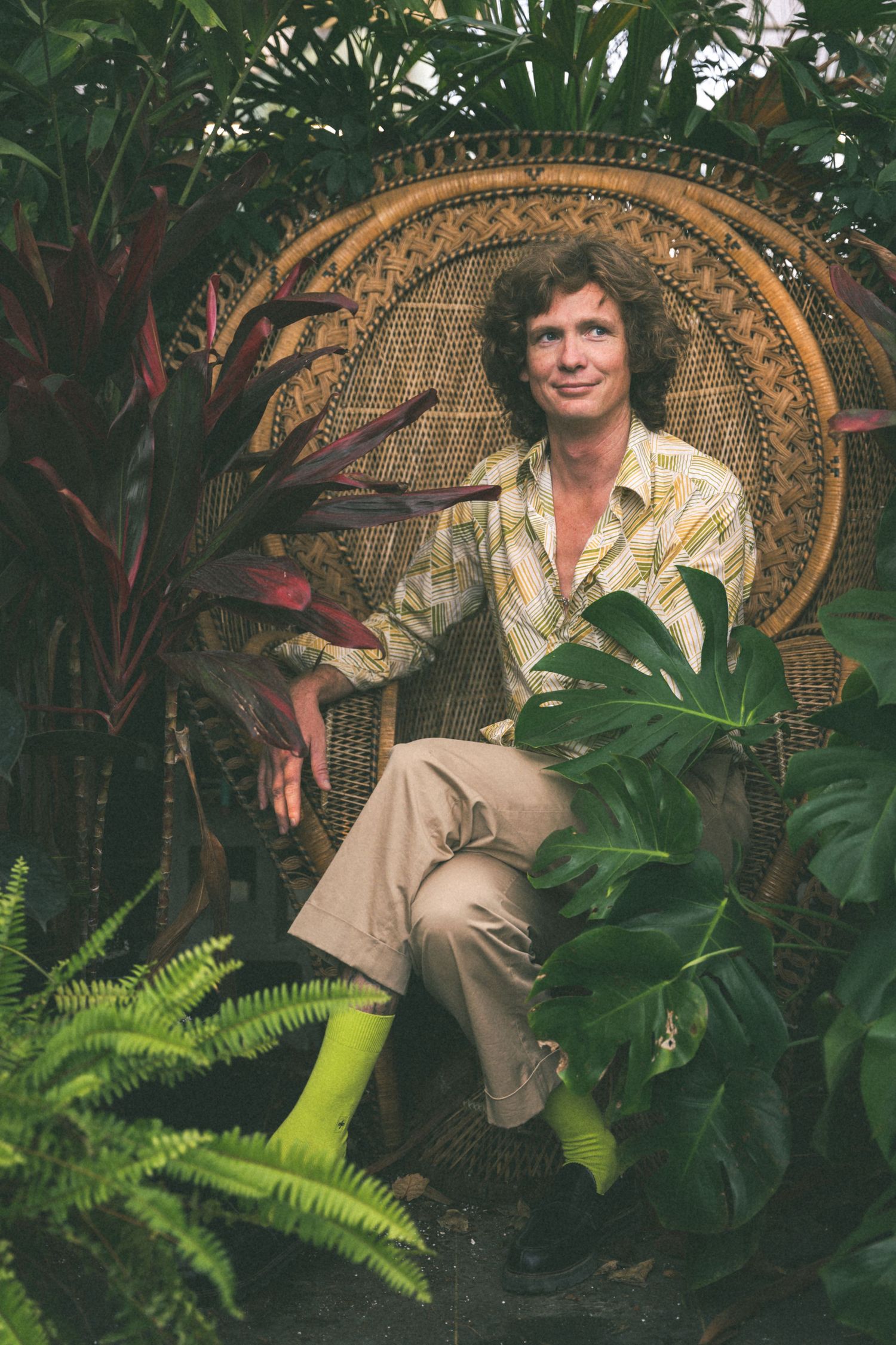 Person in floral shirt and khakis seated in wicker peacock chair surrounded by tropical plants.
