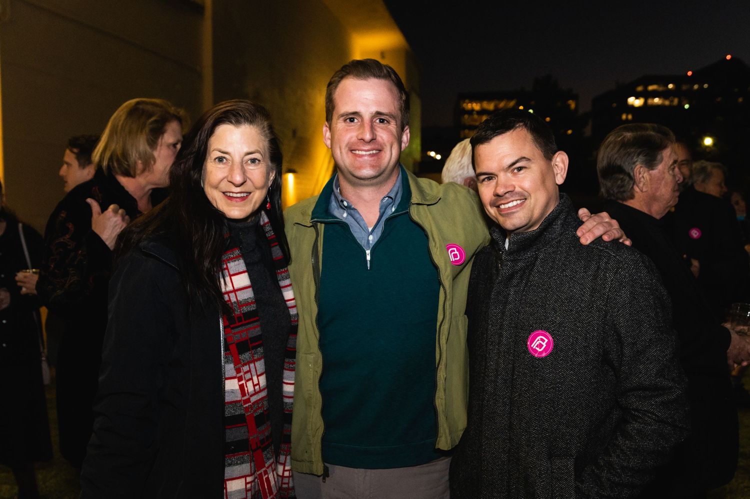 Three people in winter attire smile at evening social gathering.