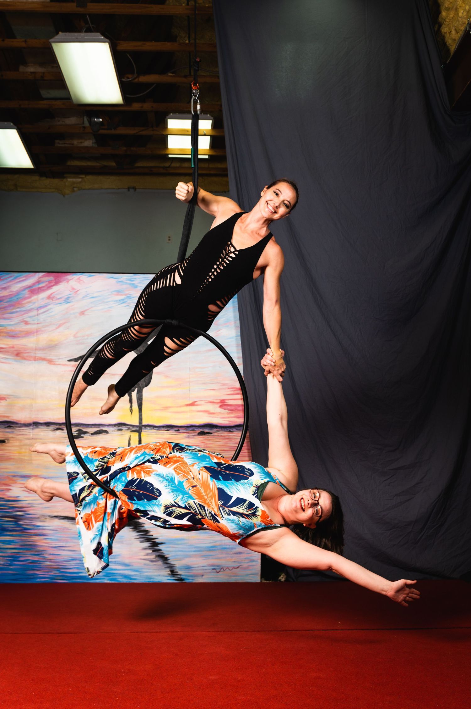 Two aerial artists perform dynamic duo moves on lyra with sunset backdrop.