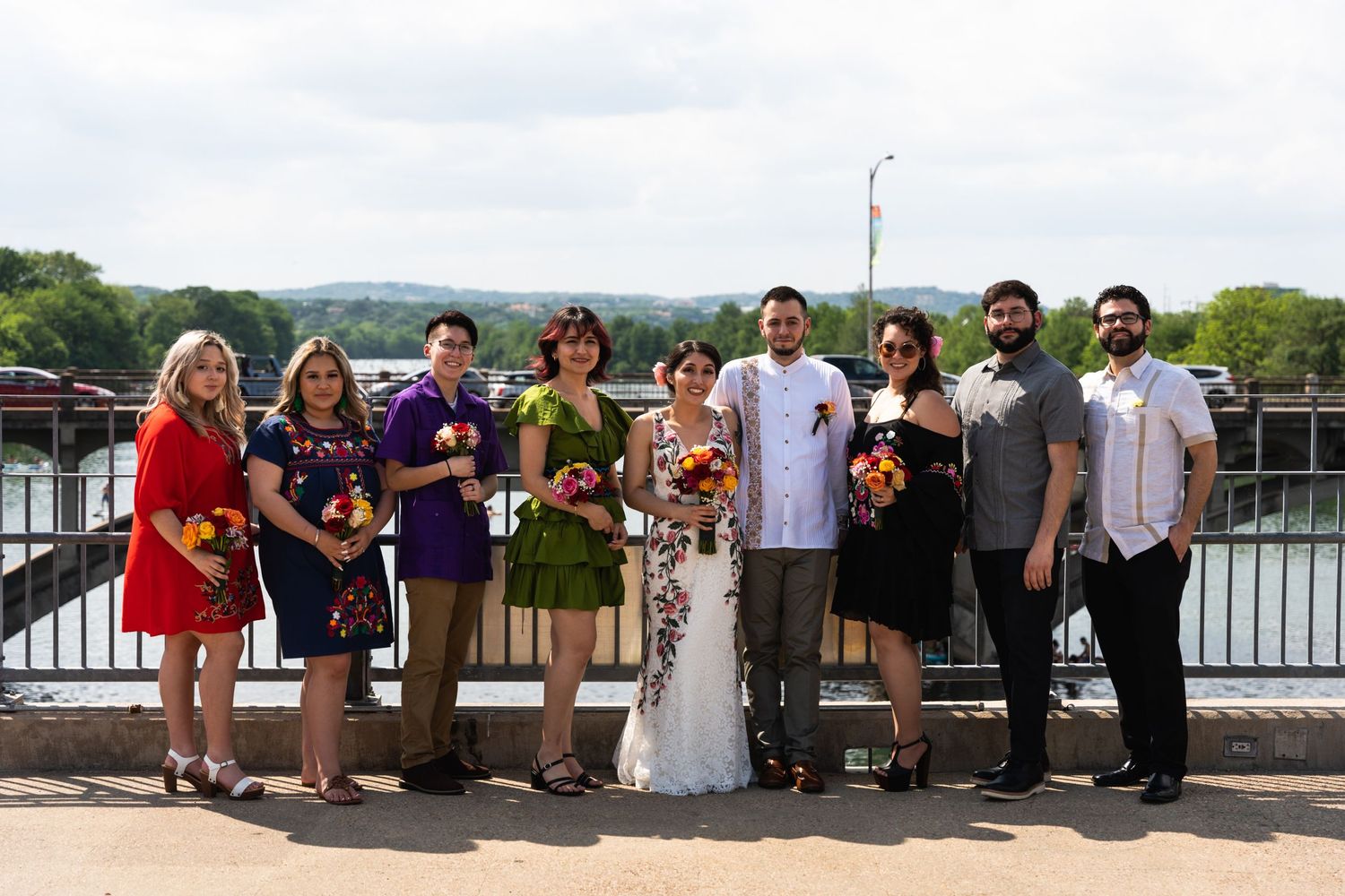 A wedding party poses together on an outdoor deck overlooking water with colorful bouquets.