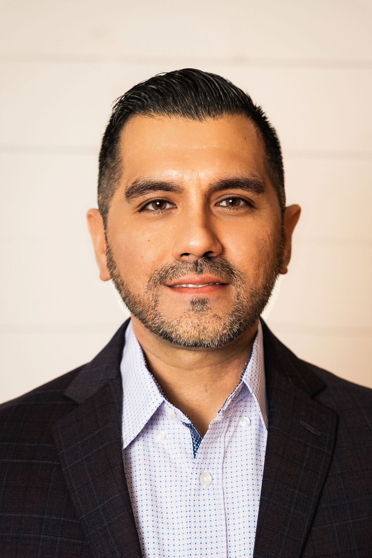 Professional headshot of person in dark suit and light blue button-down shirt against neutral background.