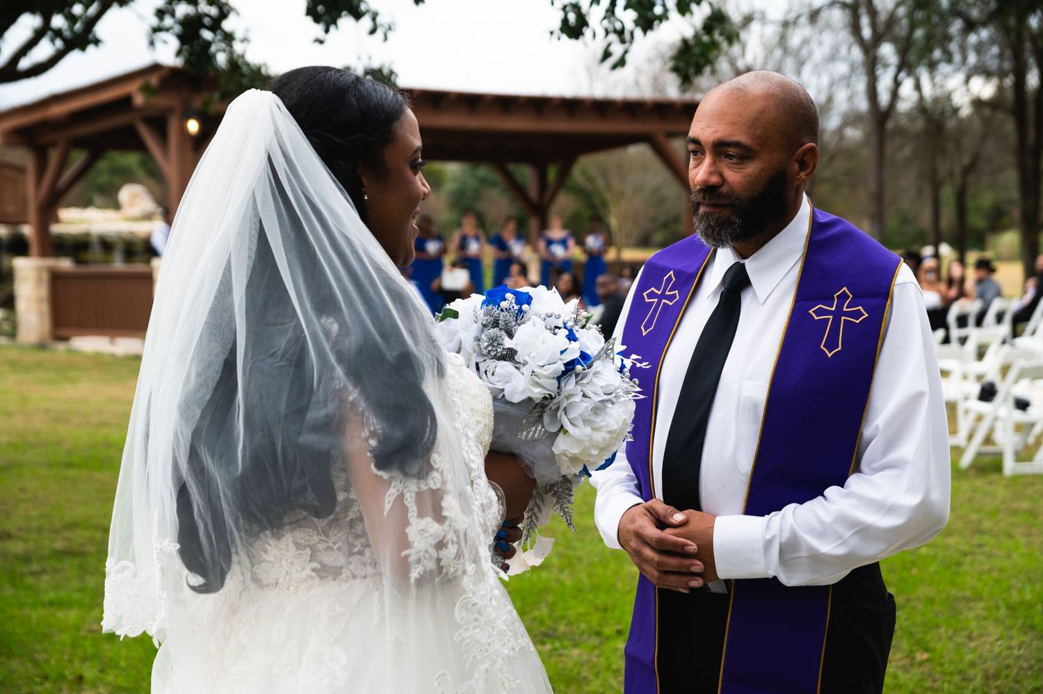 A wedding ceremony moment with the officiant in purple robes speaking to the bride outdoors.