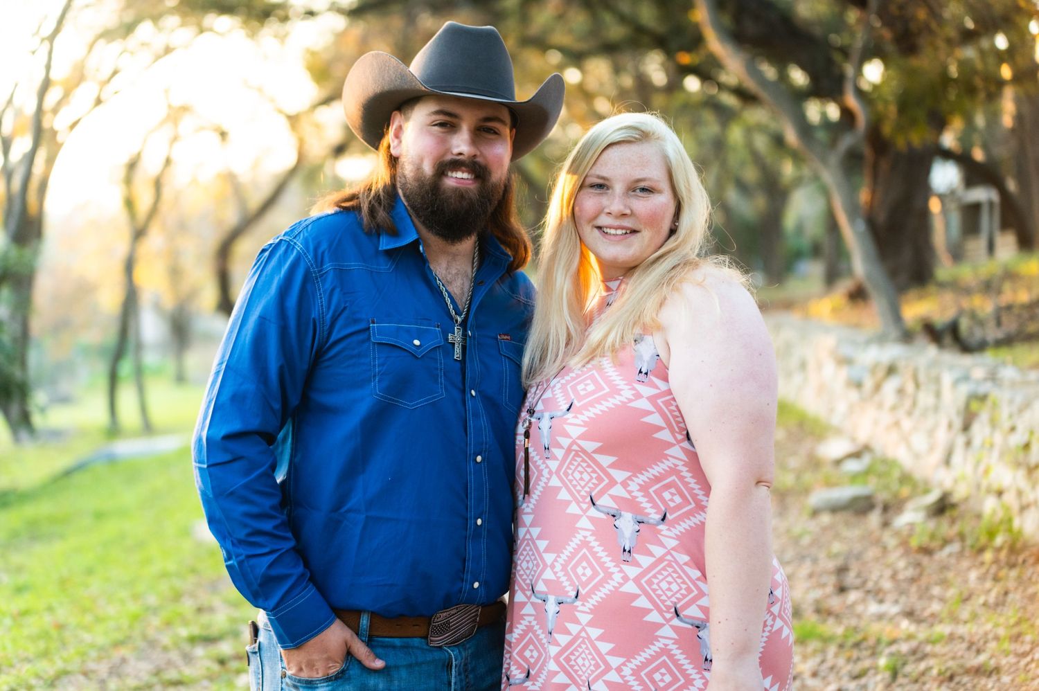 A couple in western-inspired outfits smiles at the camera against a natural outdoor backdrop.