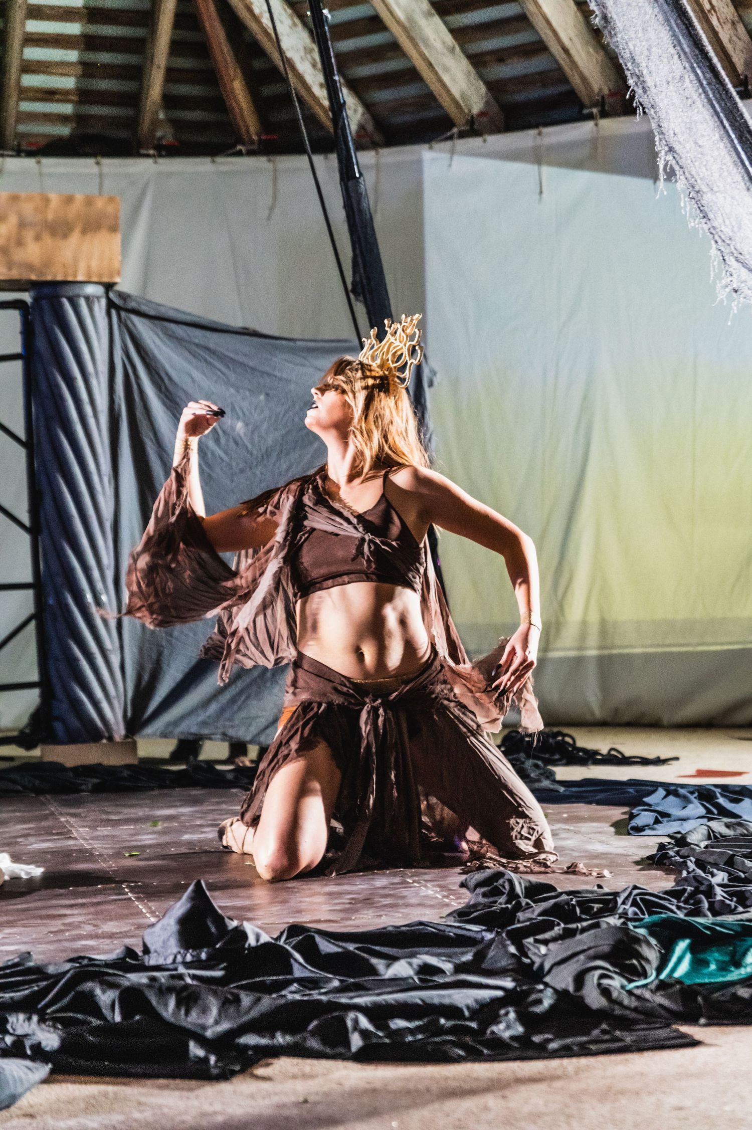 A performer in fantasy costume with crown kneels dramatically on a dark stage.